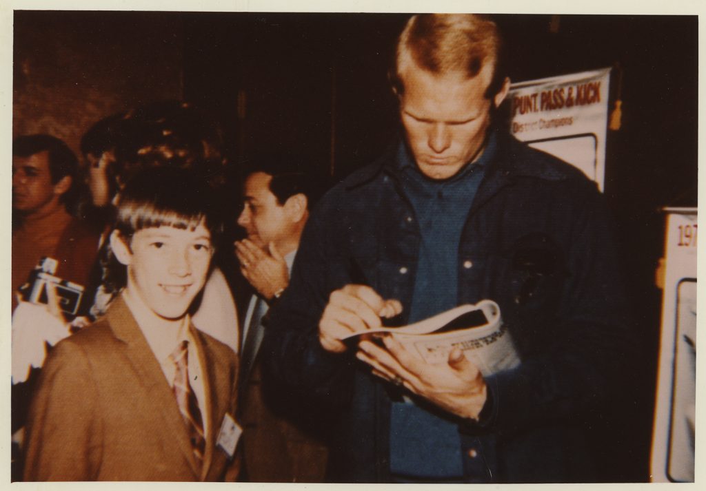 A man in a denim jacket signs a football for a boy in a suit and tie, with other people and posters visible in the museum background. *AI generated alt text