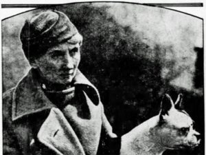 Historical black and white photo of a person in a coat and hat standing beside a light-colored dog with pointed ears; vintage look. *AI generated alt text