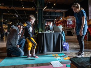 A young boy stands on a mat with colorful cards as a man kneels and a woman smiles near vintage vehicles in a museum exhibit space. *AI generated alt text