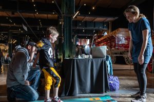 A young boy stands on a mat with colorful cards as a man kneels and a woman smiles near vintage vehicles in a museum exhibit space. *AI generated alt text