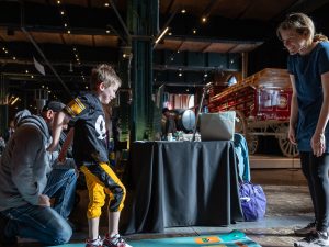 A young boy stands on a mat with colorful cards as a man kneels and a woman smiles near vintage vehicles in a museum exhibit space. *AI generated alt text