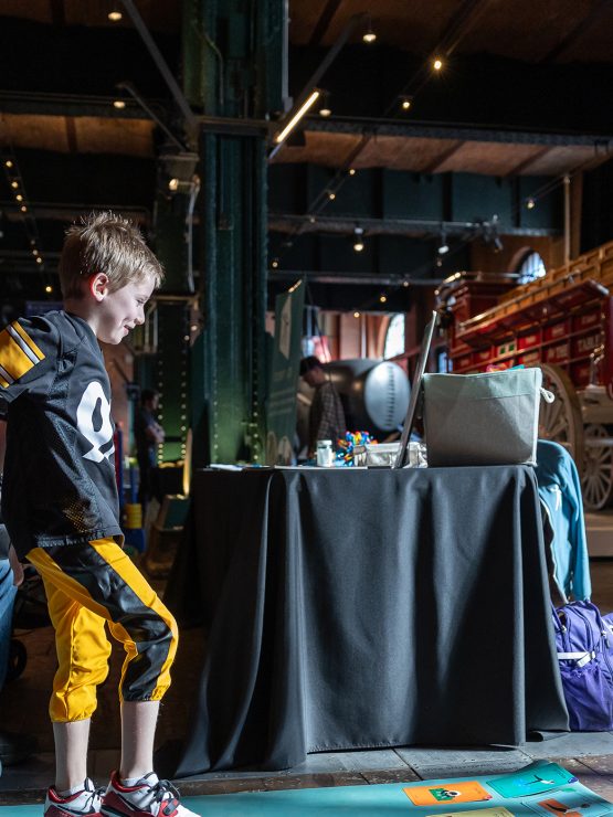 A young boy stands on a mat with colorful cards as a man kneels and a woman smiles near vintage vehicles in a museum exhibit space. *AI generated alt text