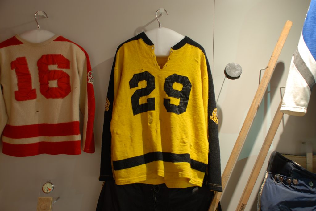 A vintage yellow and black hockey jersey with number 29 and a red and white jersey with number 16 are displayed with gear on a wall. *AI generated alt text