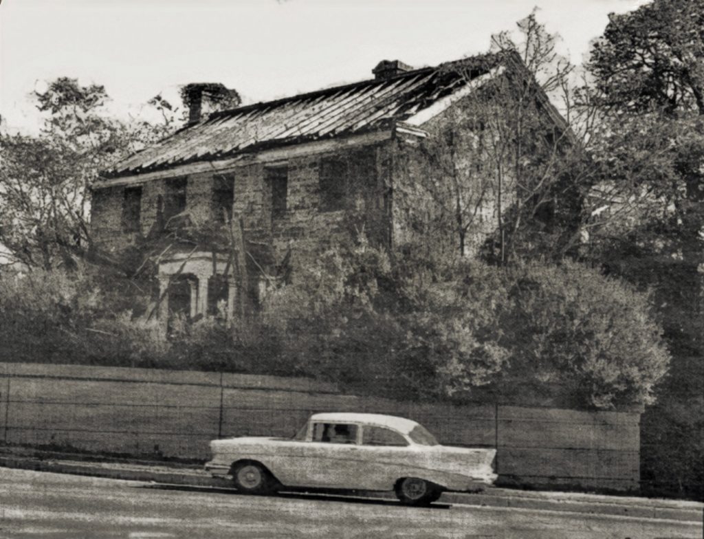 Black and white photo of an abandoned house with a damaged roof, overgrown plants, and a vintage car passing by on the street. *AI generated alt text