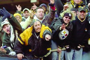 Sports fans in winter clothing and Pittsburgh Steelers gear cheer, wave, and reach out behind a barrier at a stadium. *AI generated alt text