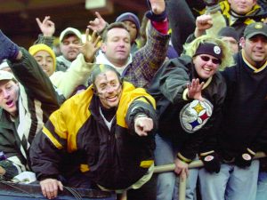 Sports fans in winter clothing and Pittsburgh Steelers gear cheer, wave, and reach out behind a barrier at a stadium. *AI generated alt text