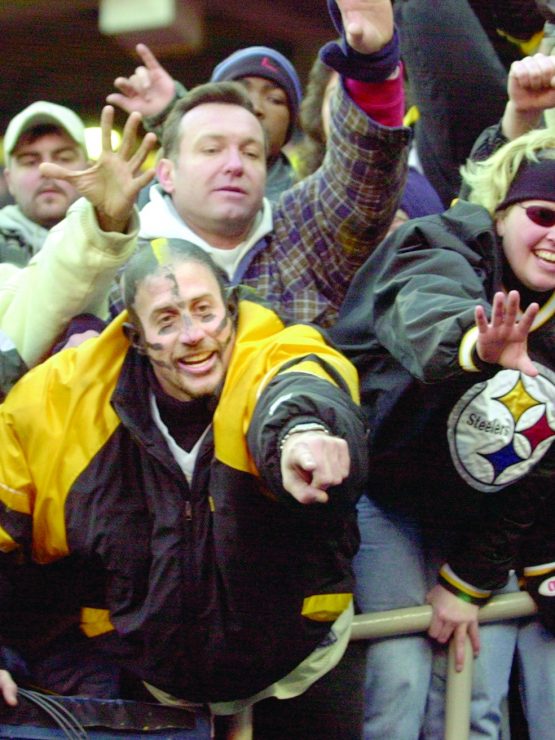 Sports fans in winter clothing and Pittsburgh Steelers gear cheer, wave, and reach out behind a barrier at a stadium. *AI generated alt text