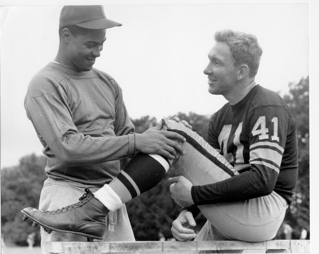 Two football players outdoors: one in a number 41 jersey sits on a bench, while another in a cap holds his leg. Both smile. *AI generated alt text