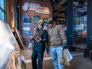 Two visitors in winter attire read a brochure at a Pennsylvania museum, with posters, shopping bags, and a large state sign behind them. *AI generated alt text