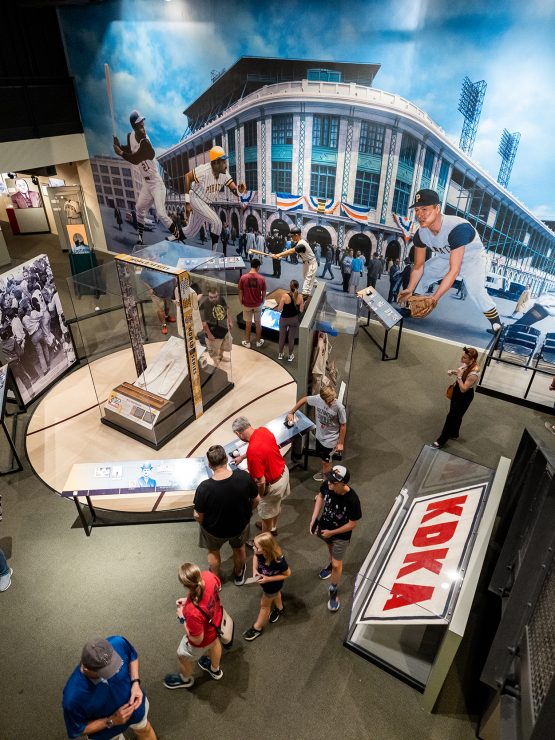 A group views a history museum exhibit with historic artifacts, display cases, old photos, murals of people in action, and a stadium backdrop. *AI generated alt text
