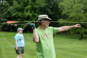 A man in a green t-shirt and hat uses an atlatl to throw a spear-like object outdoors, with another person on a grassy field. *AI generated alt text