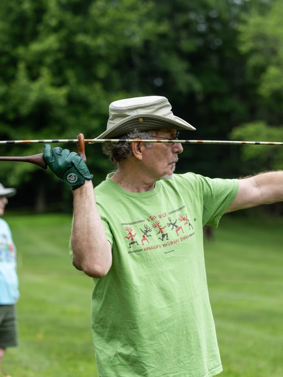 A man in a green t-shirt and hat uses an atlatl to throw a spear-like object outdoors, with another person on a grassy field. *AI generated alt text