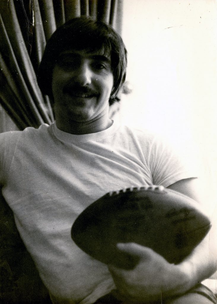 A man with dark hair and a mustache sits indoors, smiling and holding an American football. He wears a white t-shirt; curtain behind. *AI generated alt text