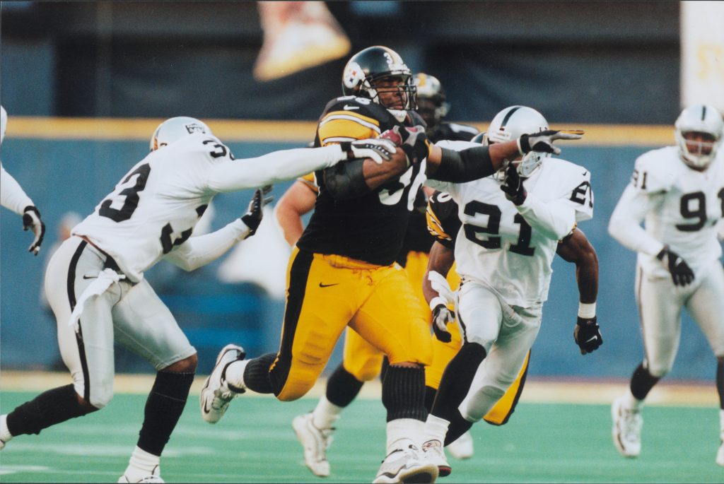 A football player in black and yellow runs with the ball, evading defenders in white on a green field during a game. *AI generated alt text