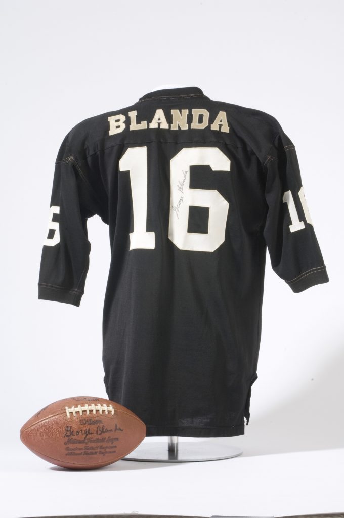 A black football jersey labeled BLANDA and number 16 in white, on display with an autographed football placed in front of it. *AI generated alt text