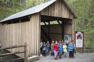 Children and adults exit a covered bridge among trees. A sign by the bridge reads 