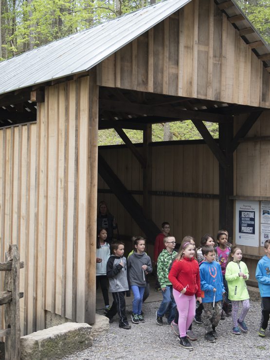 Children and adults exit a covered bridge among trees. A sign by the bridge reads 