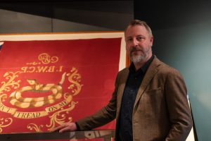 A bearded man in a brown jacket stands before a large red Fort Pitt flag with Union Jack, coiled snake, and “Don’t Tread On Me.”. *AI generated alt text