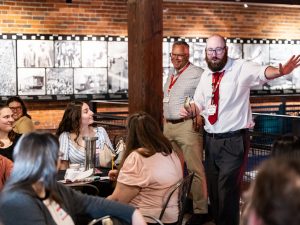 A bearded man in a white shirt and red tie speaks to a group in a brick-walled museum room with historic black and white photos. *AI generated alt text