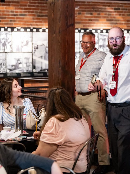 A bearded man in a white shirt and red tie speaks to a group in a brick-walled museum room with historic black and white photos. *AI generated alt text