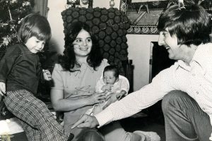 A young boy kicks a football indoors as a woman with a baby and a man sit nearby. A decorated Christmas tree stands in the background. *AI generated alt text