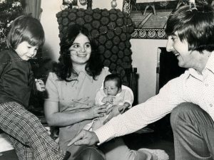 A young boy kicks a football indoors as a woman with a baby and a man sit nearby. A decorated Christmas tree stands in the background. *AI generated alt text