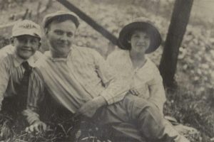 Black-and-white photo of two children and an adult seated on grass outdoors, all smiling; adult in center, each child on one side. *AI generated alt text