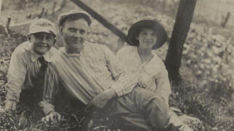 Black-and-white photo of two children and an adult seated on grass outdoors, all smiling; adult in center, each child on one side. *AI generated alt text