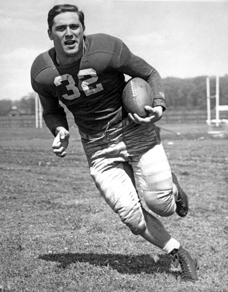 Black-and-white image of a male football player in jersey number 32 running with a football on a field, goalposts in the background. *AI generated alt text