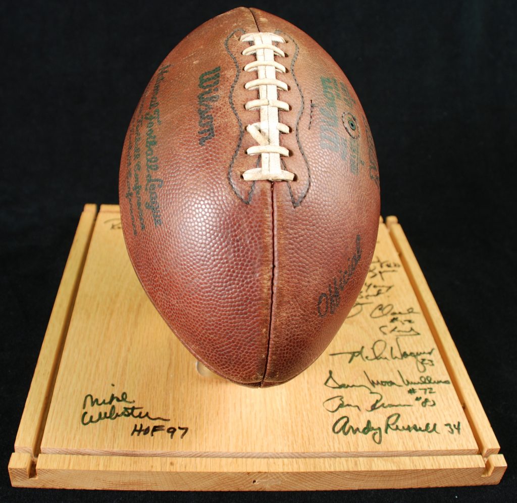 A vintage Wilson football with dark stitching on a wooden base signed by players or coaches, set against a black background. *AI generated alt text