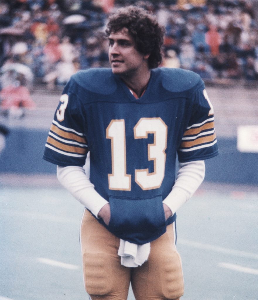 A football player in a blue jersey with yellow stripes and number 13 stands on the field; stadium seats behind are filled with spectators. *AI generated alt text