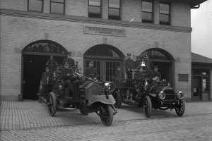 Black-and-white photo of firefighters in uniform with early 1900s fire trucks and vintage equipment outside a brick fire station. *AI generated alt text