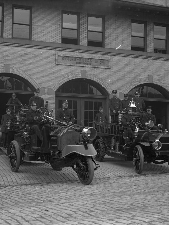 Black-and-white photo of firefighters in uniform with early 1900s fire trucks and vintage equipment outside a brick fire station. *AI generated alt text