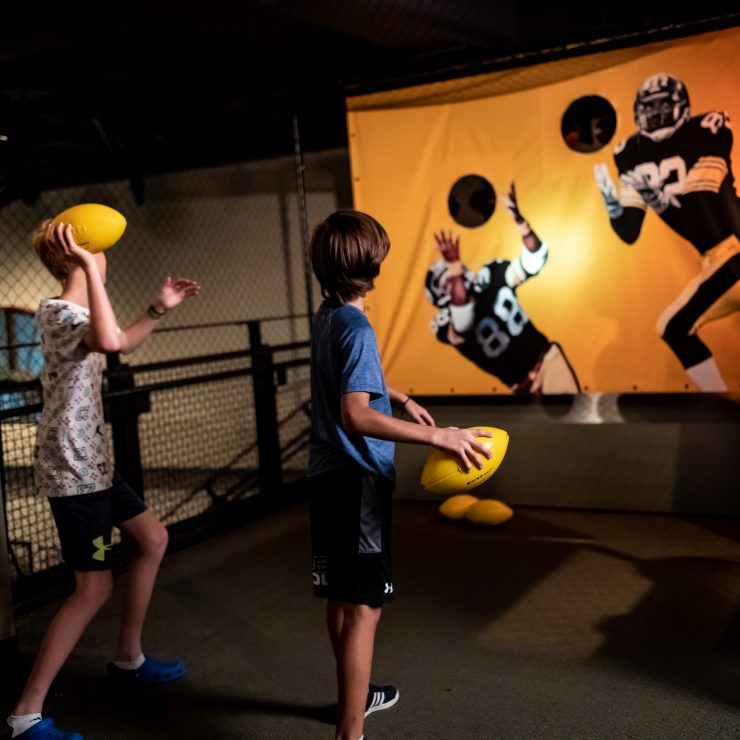 Two boys indoors aim yellow footballs at a museum display of NFL Draft players with cut-out holes as target areas on the wall. *AI generated alt text