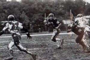 A quarterback runs on a muddy field, ready to throw, as a defender nears and two players block. Trees and a fence are behind them. *AI generated alt text