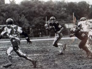 A quarterback runs on a muddy field, ready to throw, as a defender nears and two players block. Trees and a fence are behind them. *AI generated alt text