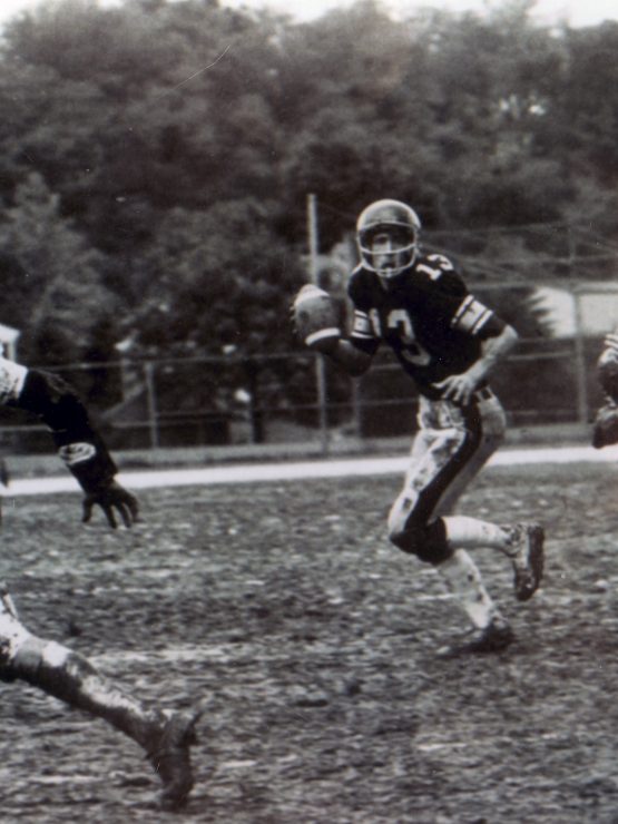 A quarterback runs on a muddy field, ready to throw, as a defender nears and two players block. Trees and a fence are behind them. *AI generated alt text