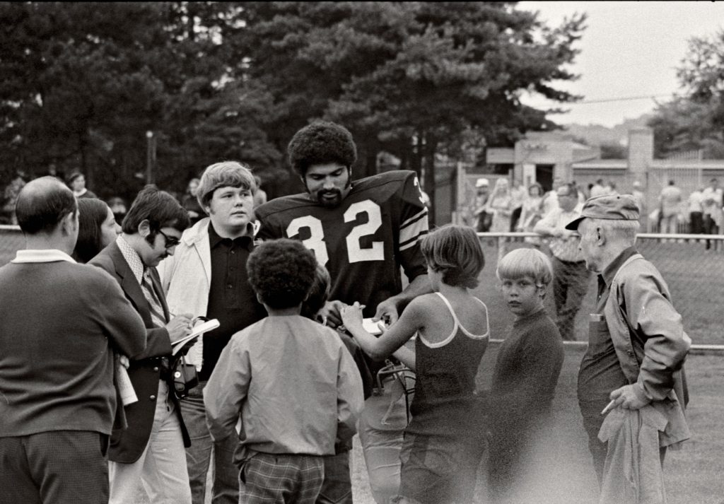 A football player wearing jersey number 32 stands outdoors with journalists, a child, and fans near a field and trees on a cloudy day. *AI generated alt text