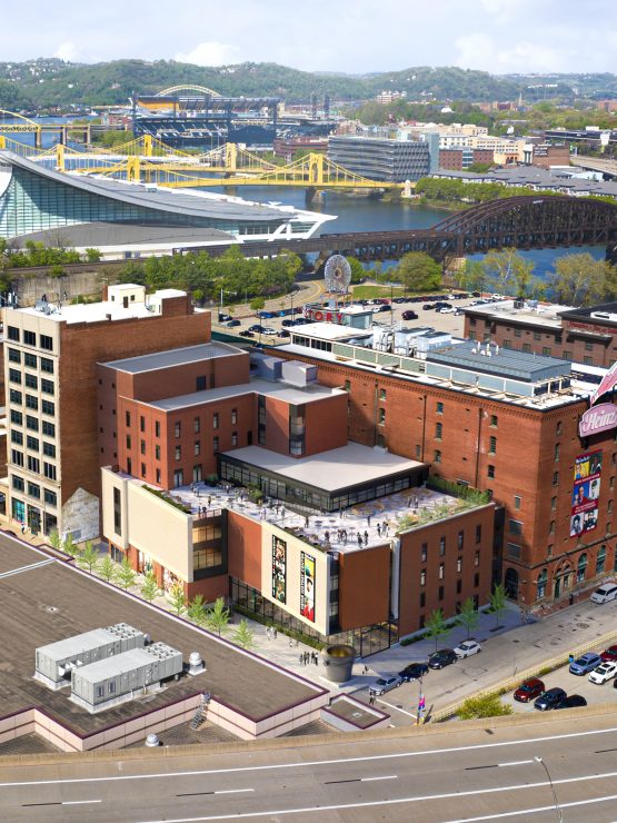 Aerial view of downtown Pittsburgh with brick buildings, rooftop terrace, river bridges, and green hills under a partly cloudy sky. *AI generated alt text