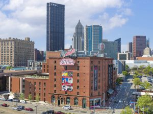 A brick building with a large Heinz sign and colorful murals in front, with tall downtown skyscrapers rising in the background. *AI generated alt text