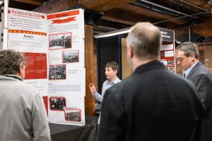 A student presents a project on the rise of the USSR to three adults in a wood-beamed room, with a large poster of text and photos. *AI generated alt text