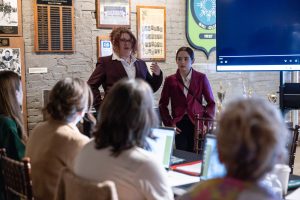 Two women in suits address a seated audience in a museum room with historic photos, plaques, and a monitor on the wall. *AI generated alt text