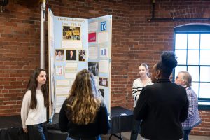Four youths and an adult stand by a “The Revolutionary Law: Title IX” board with text, photos, and charts in a brick-walled room. *AI generated alt text