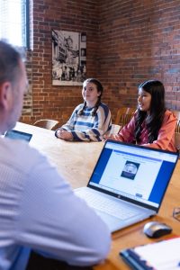 Three people meet at a wooden table in a brick-walled room; a laptop displays a webpage. A black-and-white photo hangs on the wall. *AI generated alt text