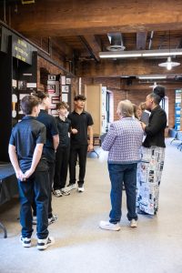 Four boys in black outfits present a project to three adults in a museum hallway with brick walls, wooden beams, and display boards. *AI generated alt text