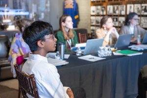 A boy in a white shirt and glasses sits in the foreground; four adults at a table with laptops and papers meet in the background. *AI generated alt text