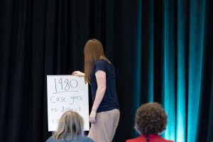 A person stands by a flip chart labeled 1980 Case goes to district co, presenting in a dimly lit room with black curtains. *AI generated alt text