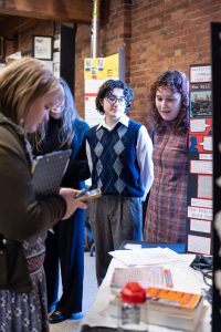 Three students stand by a project display while a woman with a notebook examines materials; exposed brick walls and posters in background. *AI generated alt text
