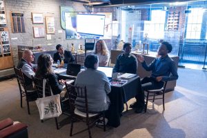Seven people sit at a rectangular table in a history museum, discussing with laptops and papers. Exhibits and plaques are in the background. *AI generated alt text