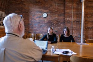 A man with white hair and glasses sits at a table with a laptop, papers, and water bottle, facing two young women in a museum room. *AI generated alt text